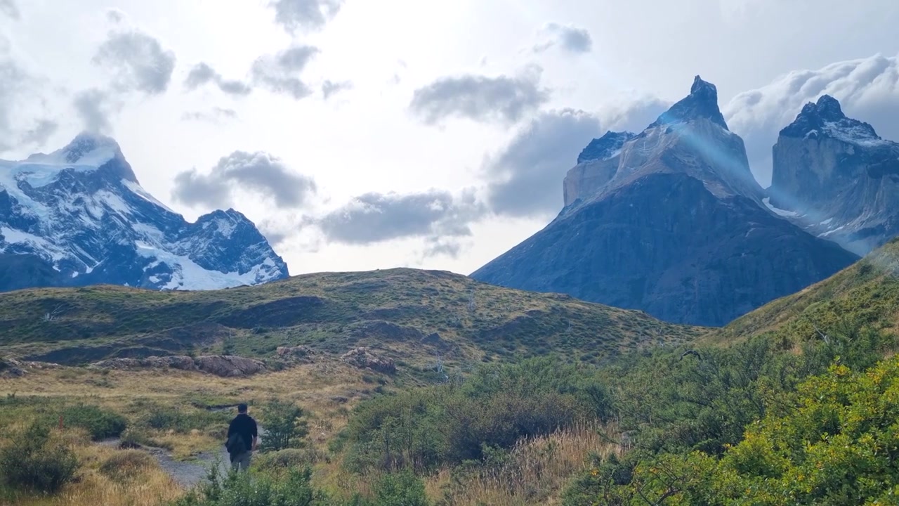 I Cuernos del Paine si ergono sopra la vasta natura selvaggia del Parco Nazionale Torres del Paine in Cile, mentre un viaggiatore solitario cammina attraverso il drammatico paesaggio patagonico sotto cieli nuvolosi.