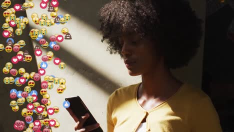 Composite-video-of-multiple-face-emojis-floating-against-african-american-woman-using-smartphone