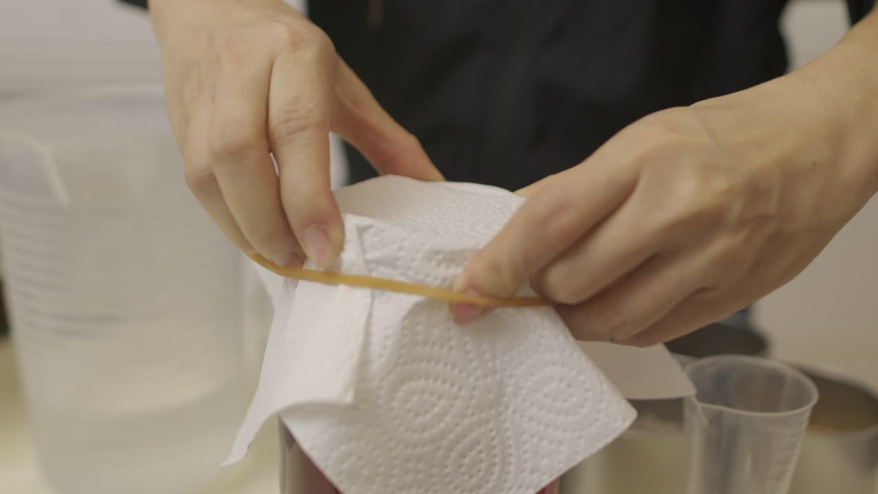 la mujer pone una cubierta de tejido en un frasco de vidrio con ingredientes de kombucha y la cierra con una banda de goma elástica