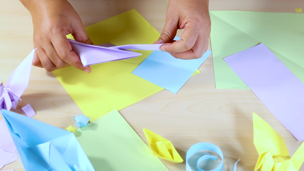 Person folds pastel origami paper, surrounded by completed models, under bright, even lighting, overhead view