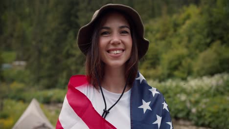 A-happy-brunette-girl-in-a-green-hat-wrapped-herself-in-the-flag-of-the-United-States-of-America-smiles-and-looks-at-the-camera-against-the-background-of-a-green-forest-and-a-mountain-river