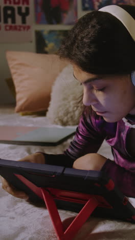 Beautiful-teen-in-headphones-lies-on-bed-in-her-room-and-uses-digital-tablet-computer.-African-American-girl-looks-at-tablet-display-and-makes-funny-faces.-Vertical-shot