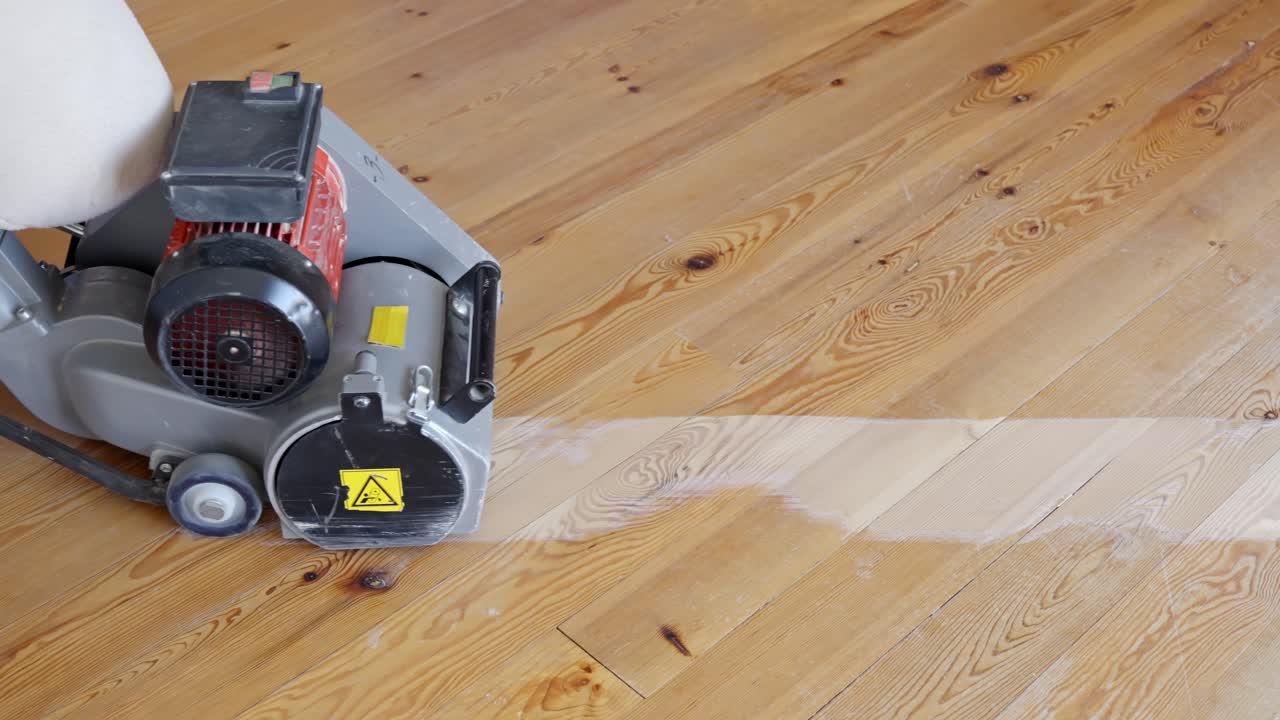 Floor sanding machine moves backward across surface leaving trail of stripped wood grain
