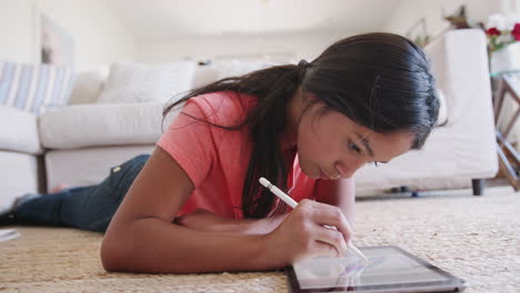 Teenage-girl-lying-on-the-floor-in-the-living-room-using-a-tablet-computer-and-stylus,-focus-on-foreground