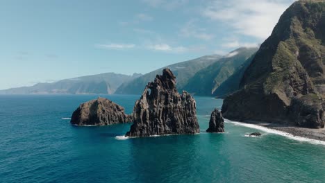 tilt-up aerial reveals the majestic sea stacks of ilhéus da ribeira da janela framed by turquoise waters and madeira’s vast green mountain range.