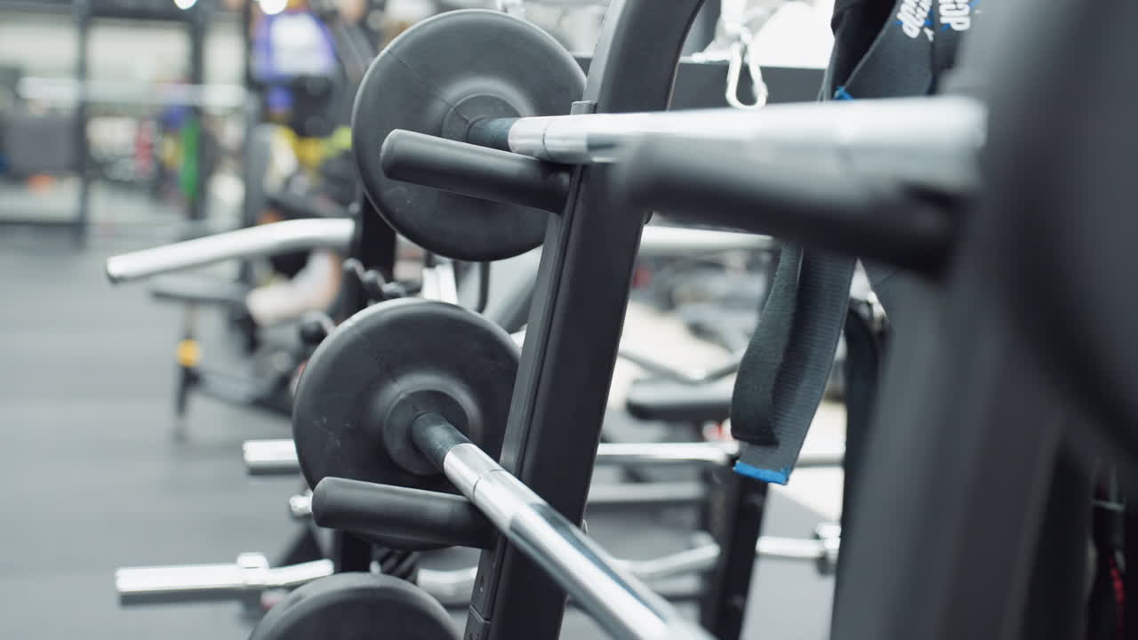 Close up of barbell loaded on machine rack, showing metal shaft, weight plates, support pegs and blurred gym interior with equipment and faint figures training, highlighting strength gear detail