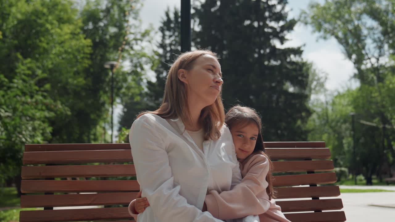 Mamma e figlia sedute su una panchina, la mamma infermiera conforta la bambina timida con un dolce abbraccio, la luce del sole filtra tra le foglie, un appoggio sulla spalla e una rassicurazione silenziosa, un momento familiare di guarigione in un parco sereno.
