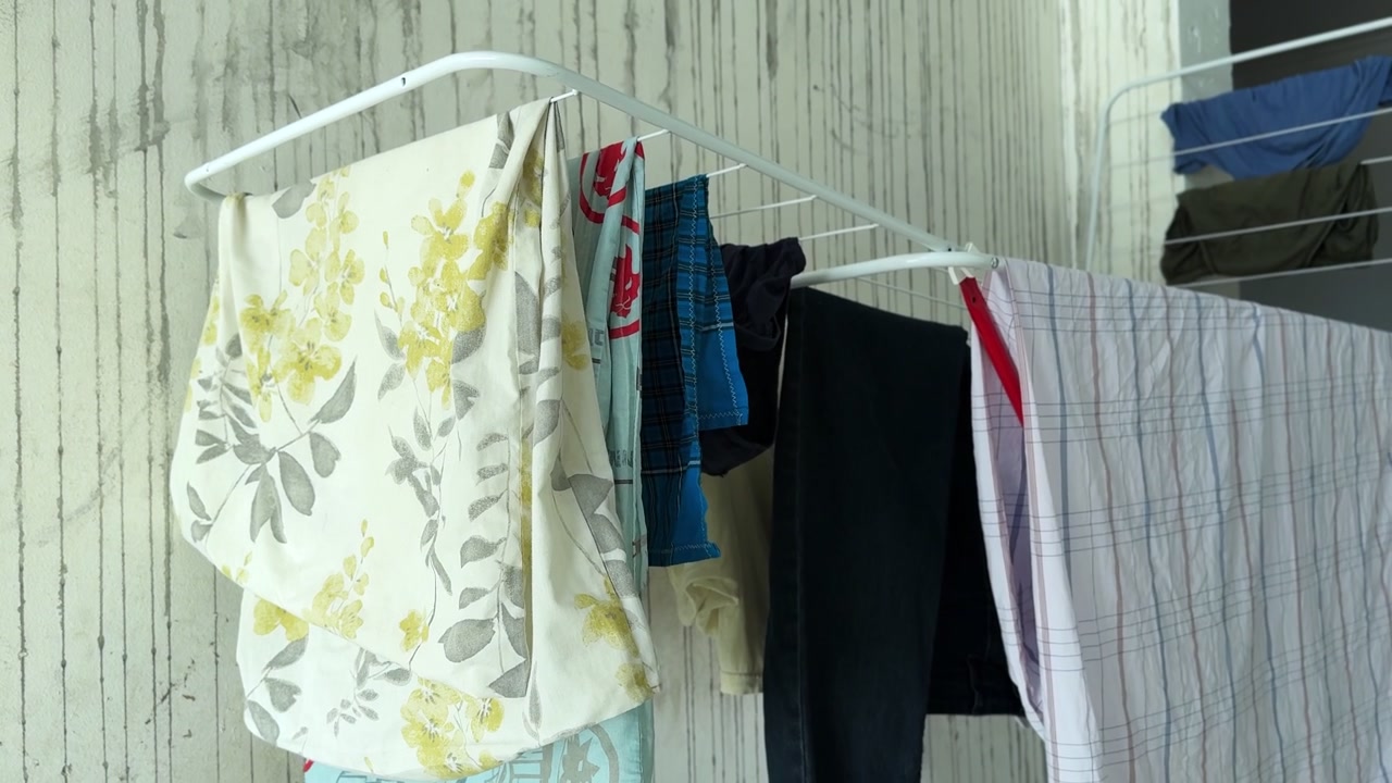 wet laundry hanging on a white metal drying rack indoors. Floral patterned fabric and dark garments air dry naturally in a brightly lit room against a textured wall during daytime.
