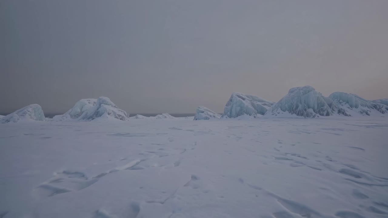Aerial video of a vast, snowy landscape with distant ice formations under a gray sky