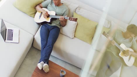 Un-Hombre-Afroamericano-Toca-La-Guitarra-Y-Canta,-Usando-Una-Computadora-Portátil-En-Casa