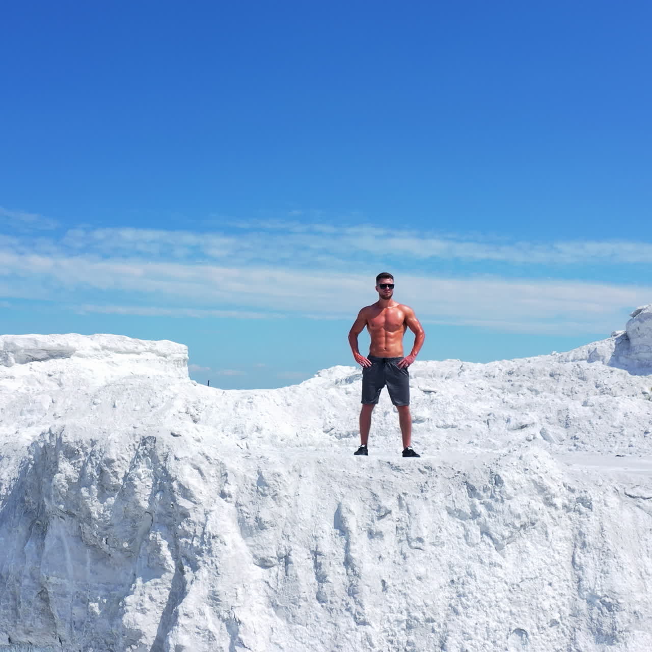 Drone view of a strong man in shorts without shirt on white hill. Young muscular man in good physical shape wearing sunglasses standing in nature. Camera moves up.