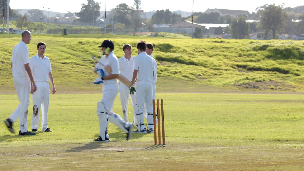 Two teams of multiracial male cricket players playing cricket, celebrating and huddling on pitch