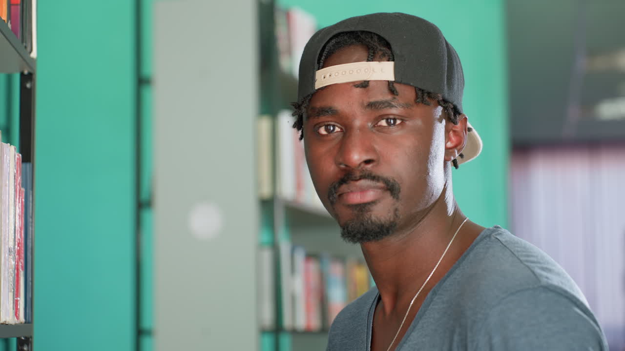 Man in cap stands near library shelf, browsing spines, then turning toward camera with quiet, focused expression, casual student style, necklace and sunglasses visible