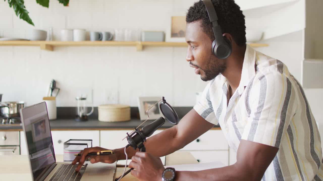 Happy african american man sitting at table in kitchen, using laptop and making vlog