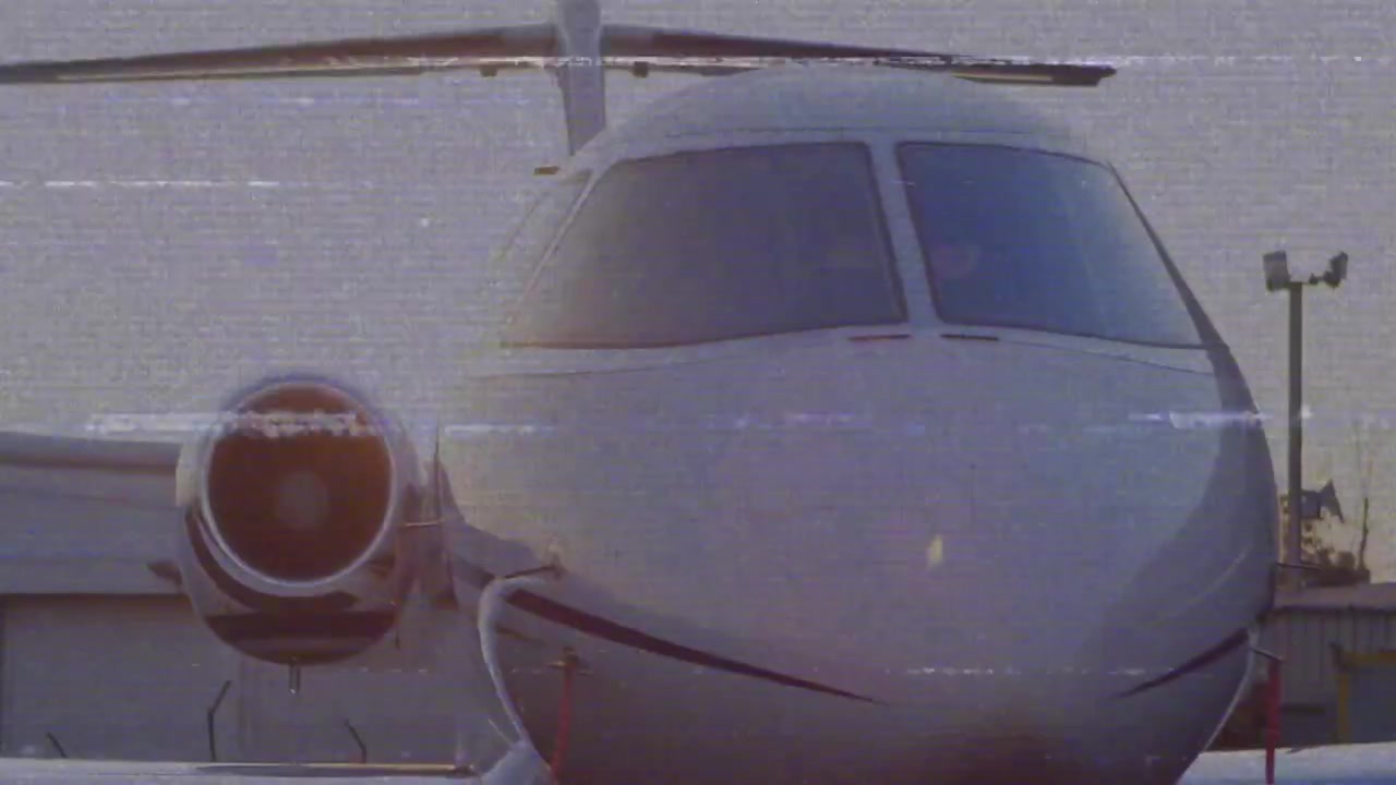Dopo un glitch statico, la telecamera zooma lentamente sul muso e sul motore di un jet, mostrando finestrini e fusoliera.