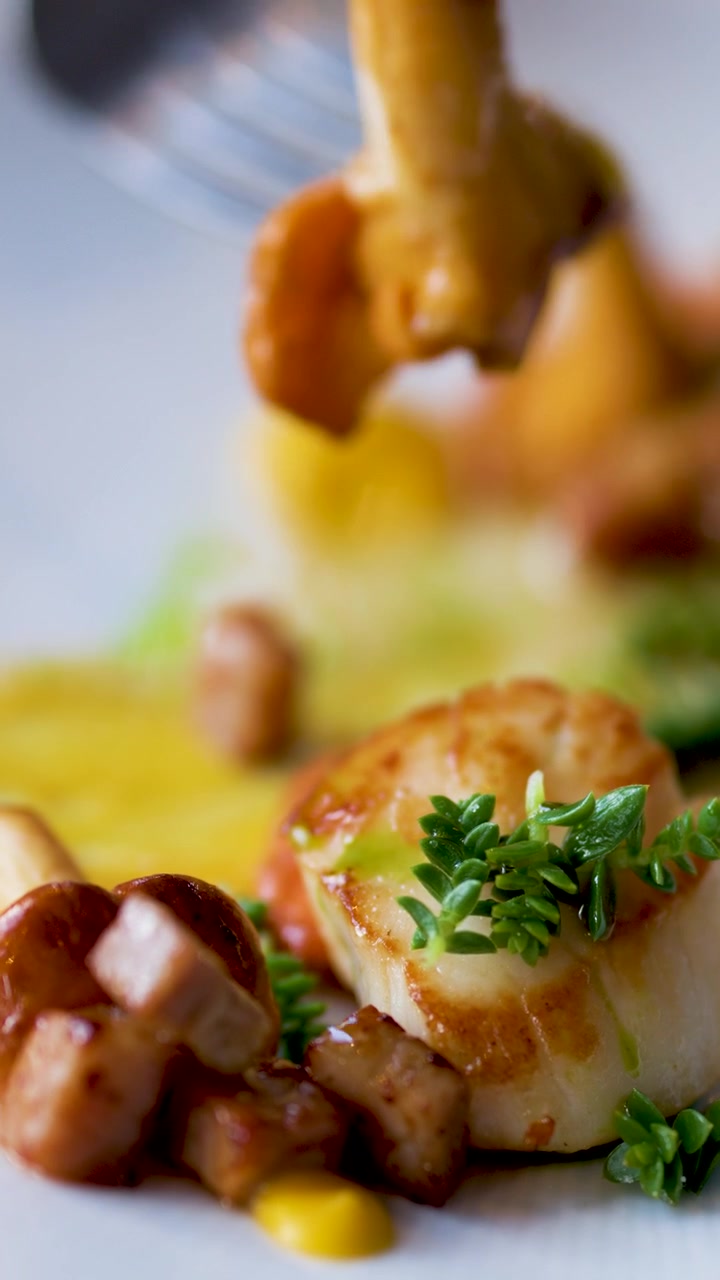 Una forchetta solleva una capasanta scottata guarnita con erbe aromatiche da un elegante piatto di frutti di mare, sotto una luce naturale soffusa, con una profondità di campo ridotta e prospettiva ravvicinata.