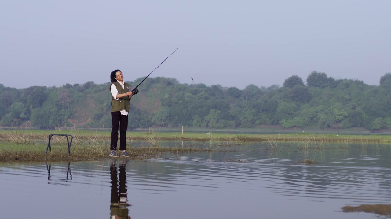 A South Asian girl wearing outdoor vest fishes quietly while standing alone in a lake, seen from behind
