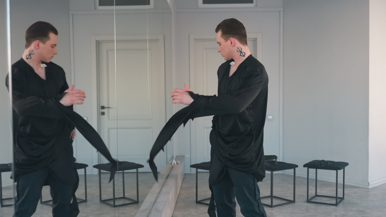 young man stands before full length mirror in bright studio, pulls hood of tattered black costume over head, revealing jagged edges and edgy silhouette for dramatic Halloween transformation