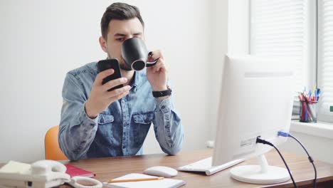 Young-office-worker-using-his-phone-at-the-office-sitting-at-the-table-with-computer,-phone-and-drinking-coffee-or-tea-from-his