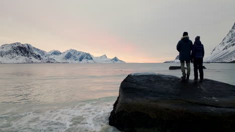 Travelers-on-rock-look-out-into-bay-surrounded-by-snowy-mountains,-sunset-aerial