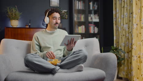 young man watching comic movie on tablet while eating chips and sitting with crossed legs on couch at home