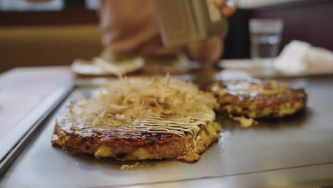 Bonito-Fish-Flakes-being-added-to-Okonomiyaki,-Japanese-Restaurant