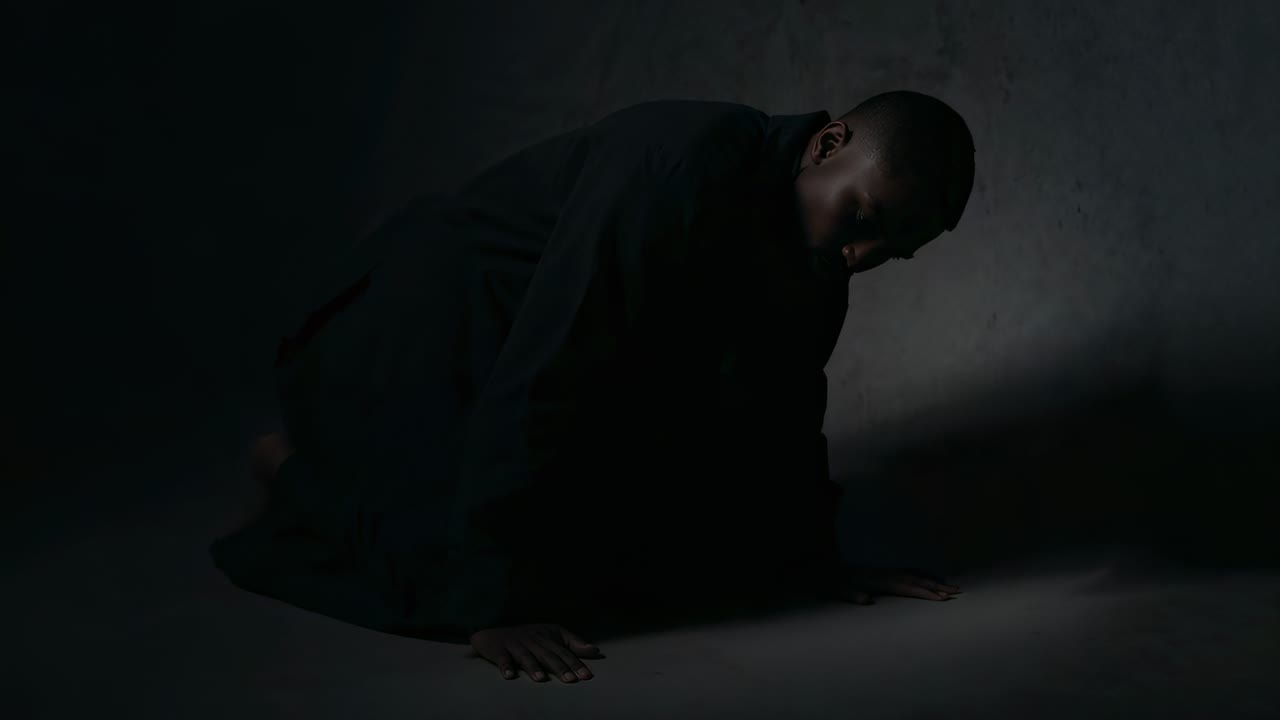 Uomo inginocchiato con cappotto scuro, palmi delle mani sul pavimento di uno studio illuminato da un fascio di luce stretto, parete ruvida