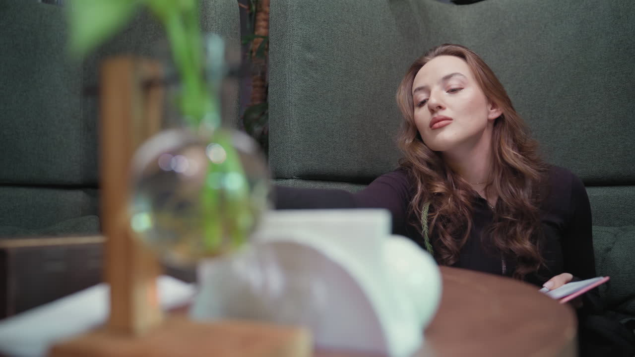 Blonde woman wearing black top sits thoughtfully in green booth holding pink notebook as she writes, with soft focus foreground with tabletop decoration and lush plants enhancing peaceful atmosphere