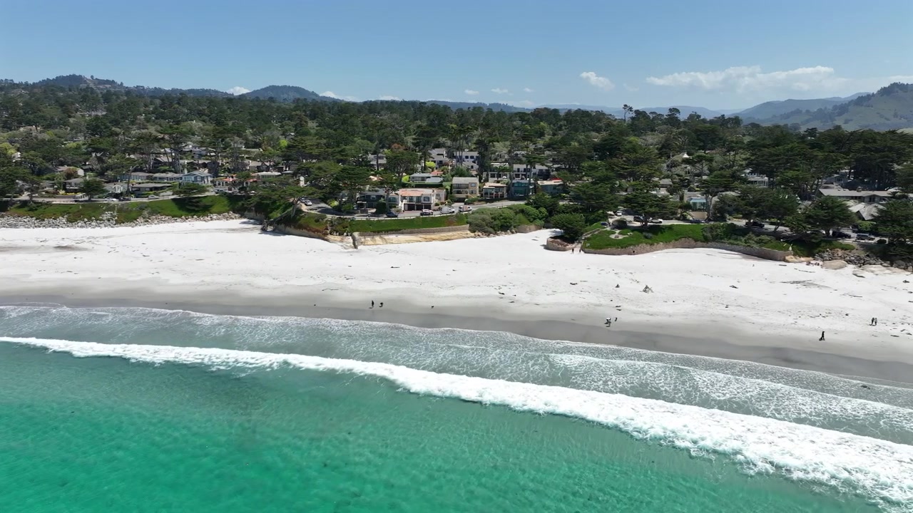 Cinematic aerial drone footage of Carmel-by-the-Sea, California, showcasing luxury coastal homes overlooking a wide white sand beach, lush green trees, scenic residential neighborhoods
