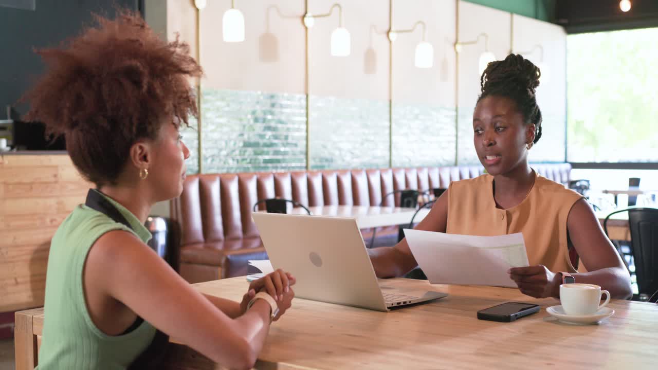 African American women talking at cafe, left woman explaining, right reading laptop papers deciding