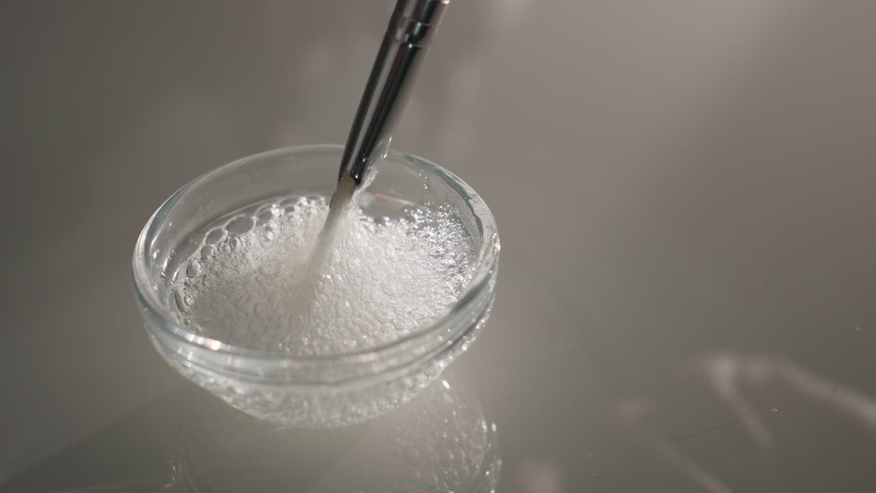 Face mask gel with bubbles lifted by gloved brush from small glass plate under soft light highlighting clear viscous texture for professional skincare treatment preparation and quality inspection