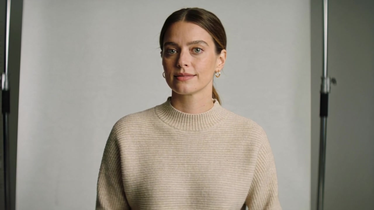 Kvinde i beige strik, der blinker, følger anvisning, drejer hovedet, lukker øjnene, med grå baggrund og lysstativer.