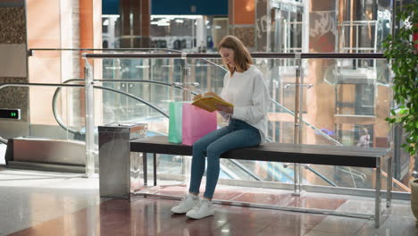 mujer joven sentada en un banco en un vibrante centro comercial leyendo un libro amarillo mientras balancea la pierna lentamente, bolsas de compras de colores visibles, barandillas de vidrio modernas y ascensor en movimiento