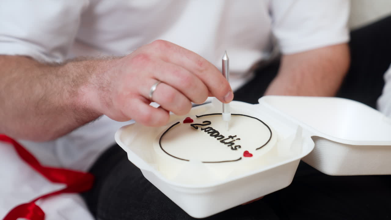 Unrecognized man holding a food box with a cake signed two months. Hand puts a candle into a cake. Close up.