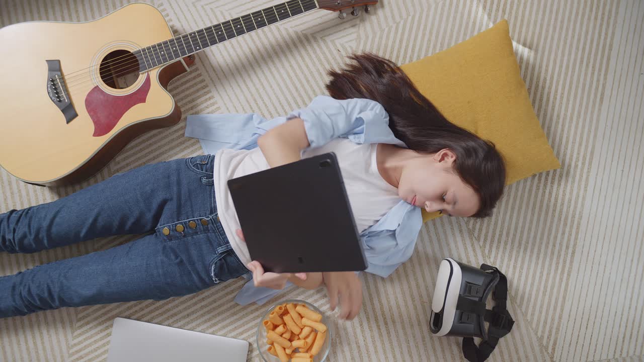 vista superior de una adolescente asiática comiendo bocadillos y usando una tableta mientras yacía en la alfombra en el suelo de su casa