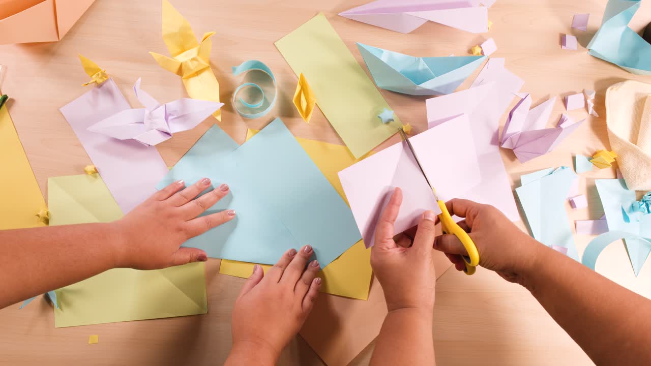 Overhead view of hands using scissors to cut paper amidst various origami creations and supplies