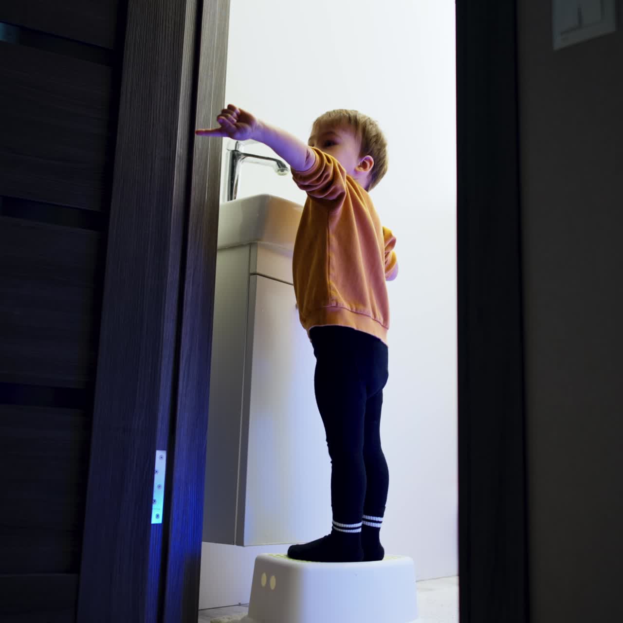 Toddler boy stands on the prop near the sink in the bathroom. Kid opens the faucet and watching the water flow. Low angle view