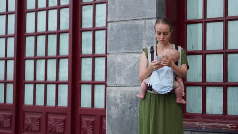 a young woman travels with her baby in a kangaroo backpack and photographs the experience with her mobile phone. as they stroll, she occasionally checks the screen