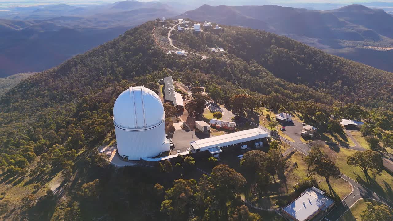 Aerial drone footage orbits a large astronomical observatory atop a forested mountain in early morning light, revealing surrounding landscape and research buildings
