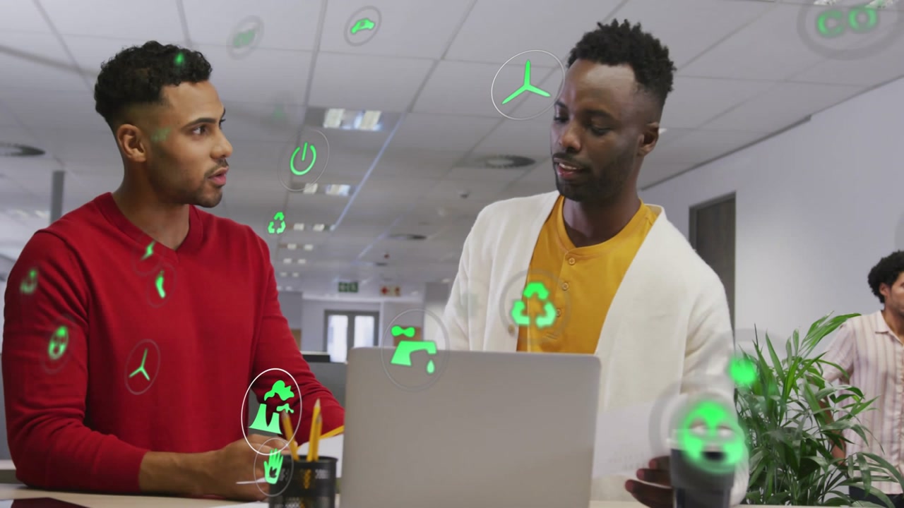 Left man pointing right man noting paper reviewing laptop icons floating amid men on office desk