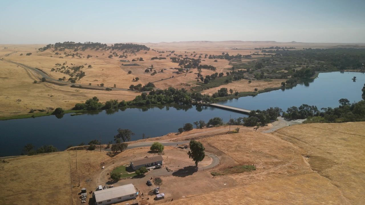 Órbita de drone em ângulo elevado sobre o Rio Merced com a Ponte Merced na Califórnia, cercado por paisagens naturais secas e serenas.