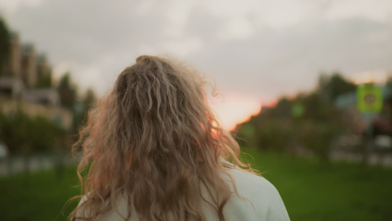 close up back view of girl in white coat walking along path twirling umbrella playfully during golden hour with curly hair swaying and blurred background showing signpost, grass, person approaching