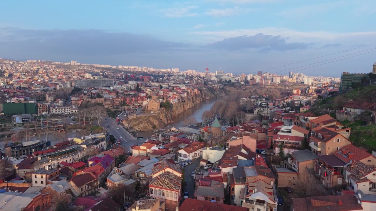 Ascesa del drone che rivela la Chiesa di Metekhi, il ponte sul fiume Kura e lo skyline di Tbilisi. Luce calda sulla capitale georgiana storica e moderna.