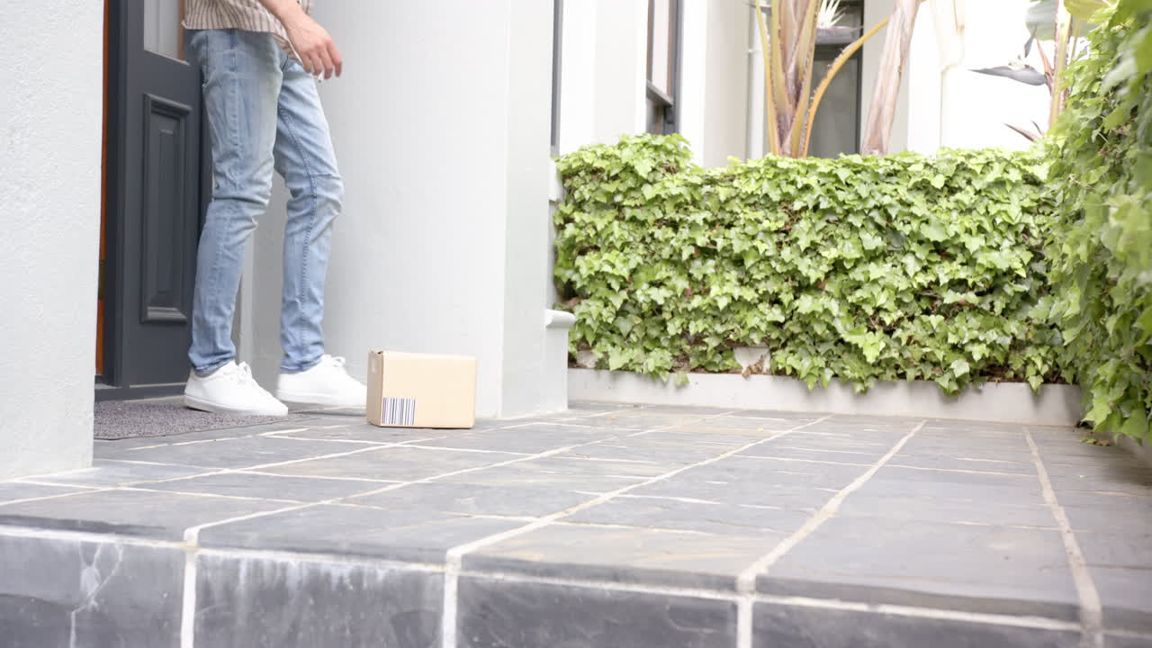 Biracial man picking up delivery in cardboard box outside front door, copy space, slow motion