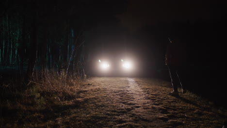 person standing on a dark forest road at night with car headlights