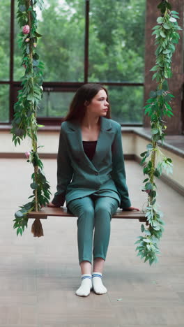 tired businesswoman sits on swings in studio. rich young woman wearing green formal suit without shoes rests after exhausting meeting. mental health