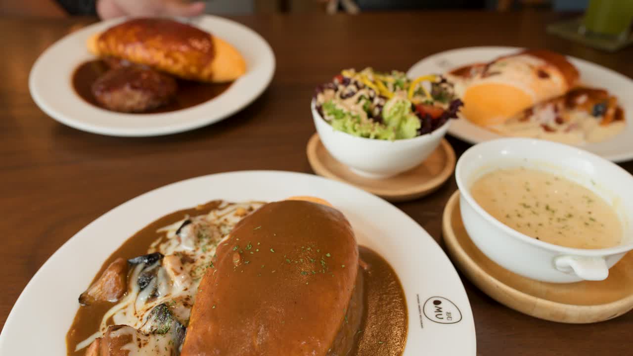 Creamy omelette rice, salad, soup, and breaded cutlet arranged on wooden restaurant table, natural lighting