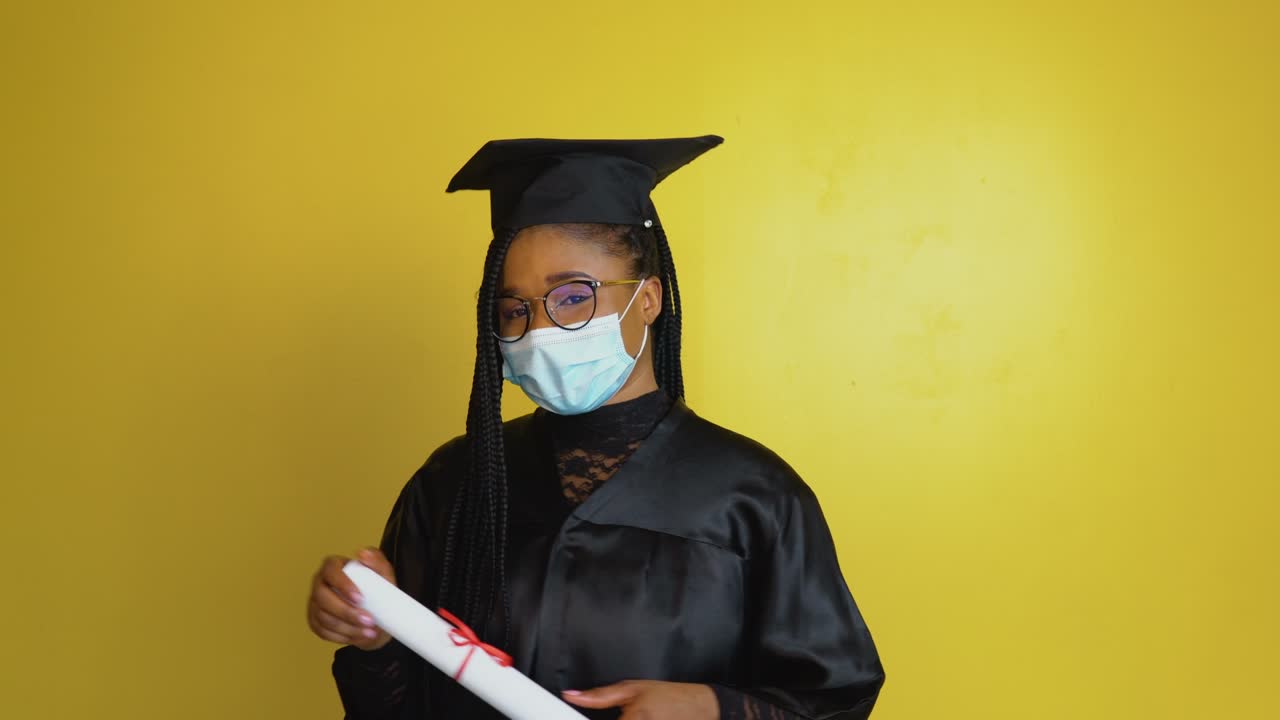 A graduate student in a protective mask points with a diploma in hand towards the camera. Student in a black robe and hat on a yellow solid background looks at the camera