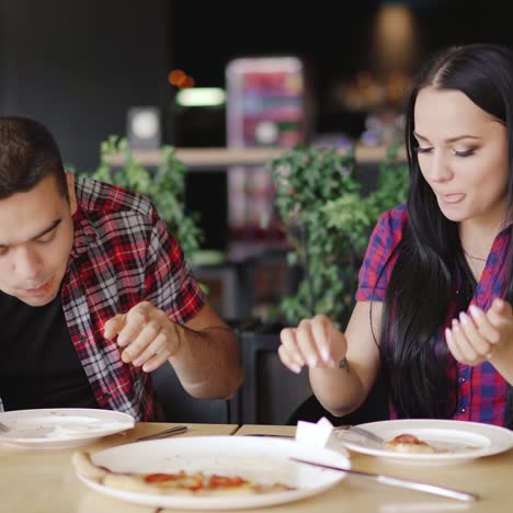 a man and a woman are eating one slice of pizza from two sides at the table in the restaurant on the background of modern interior