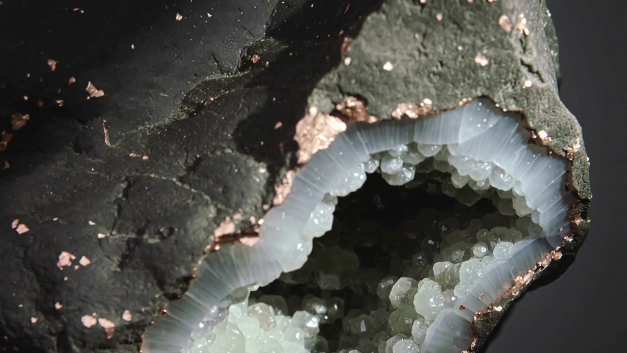 La telecamera si allontana, rivelando un geode aperto su una base scura, mettendo in evidenza cristalli druzy e scaglie di rame.